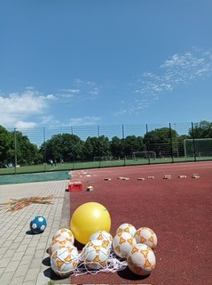 Mehrere Fußbälle und ein großer gelber Ball auf einem Sportplatz unter klarem Himmel.