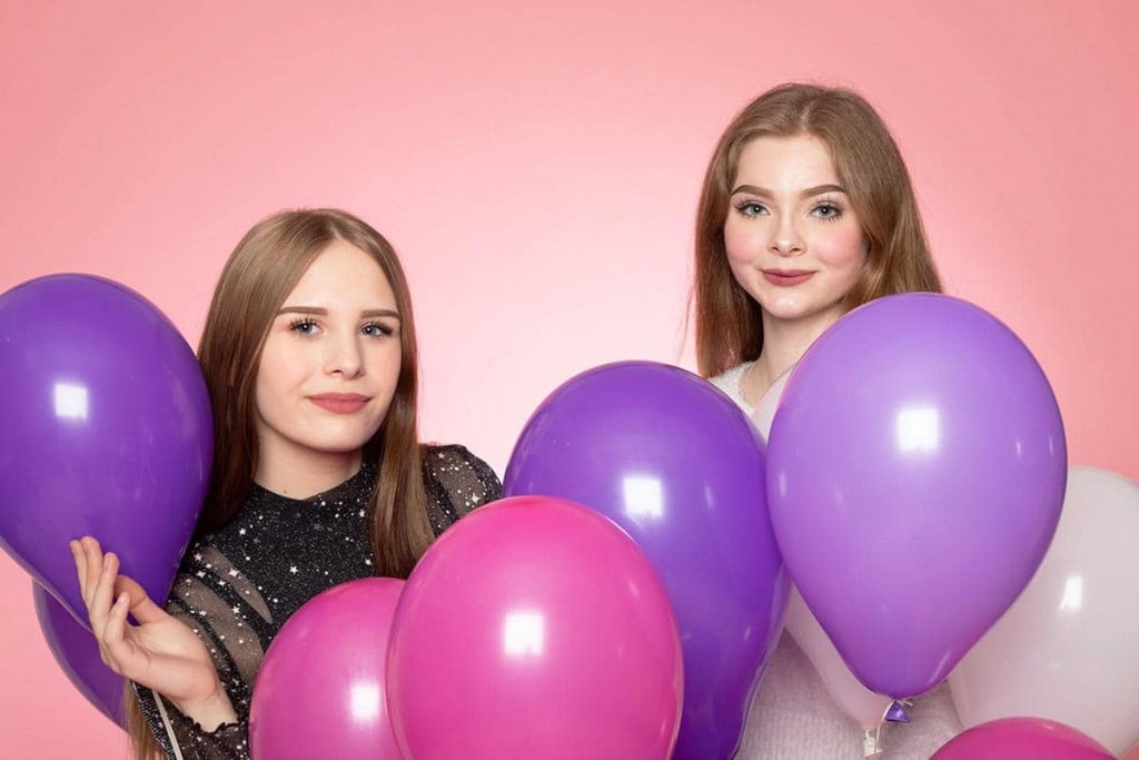 Zwei lächelnde Frauen mit braunem Haar halten lila und rosa Luftballons vor rosa Hintergrund.