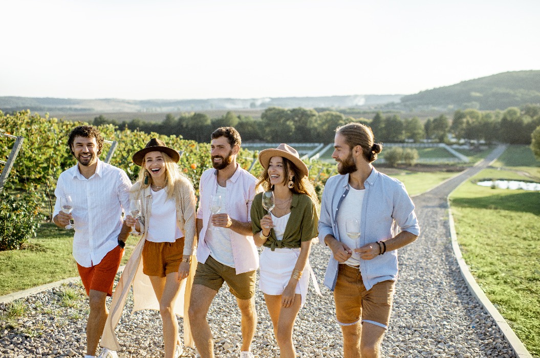 Eine Gruppe junger, leger gekleideter Freunde, die an einem sonnigen Tag mit Weingläsern auf einem Weinberg spazieren gehen