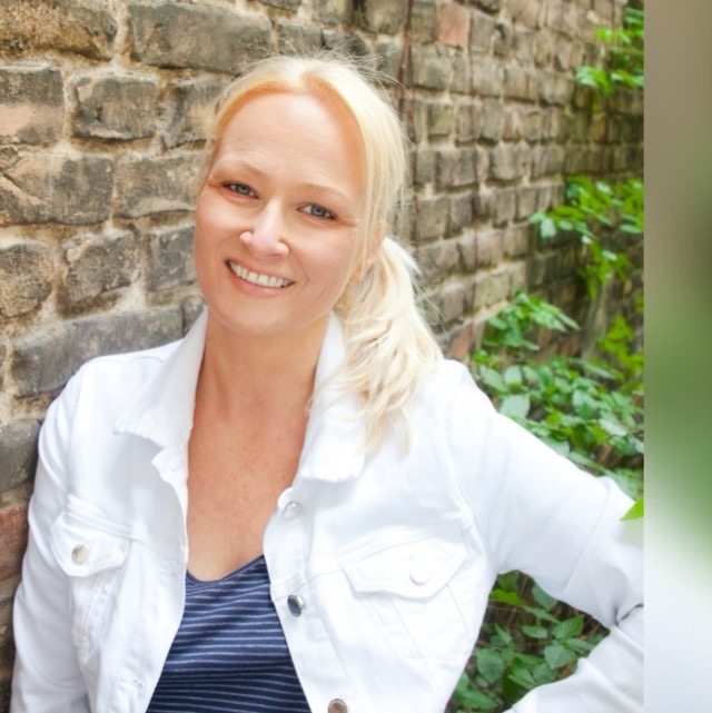 Eine lächelnde Frau mit blonder Frisur, weißer Jacke vor einer Ziegelwand mit Pflanzen.