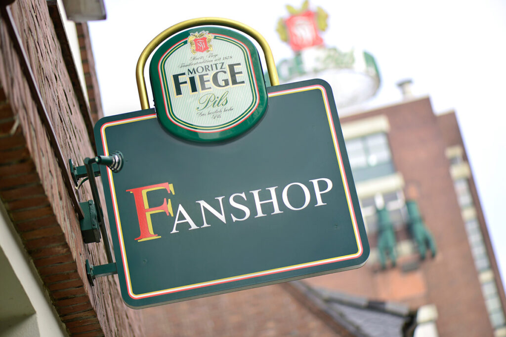 Schild mit "Fanshop"-Aufschrift, grüner Hintergrund, Brauerei-Motiv, Außenansicht.