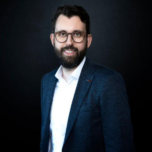 Ein Mann mit Bart und Brille lächelt vor schwarzem Hintergrund, trägt ein dunkelblaues Sakko.