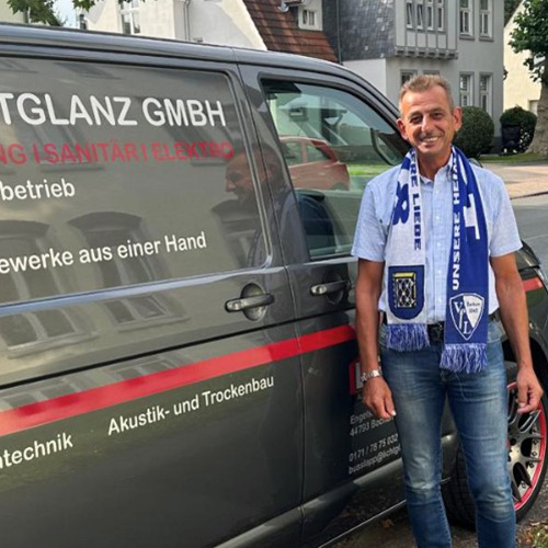 Ein gut gelaunter Mann mit Fußballschal steht vor einem Firmenfahrzeug.