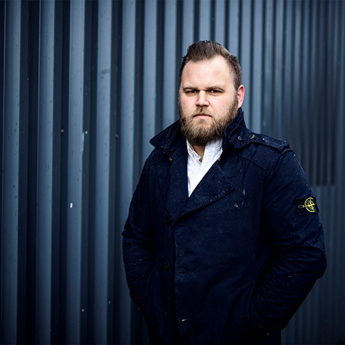 Mann mit ernstem Blick in dunkelblauer Jacke vor einer gerillten Metallwand