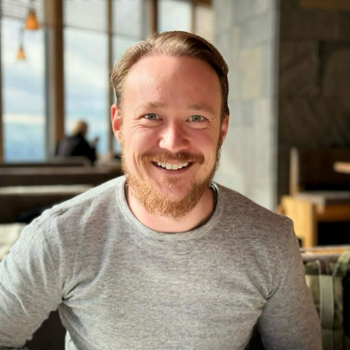Ein lächelnder Mann mit roten Haaren und Bart trägt ein graues Shirt.