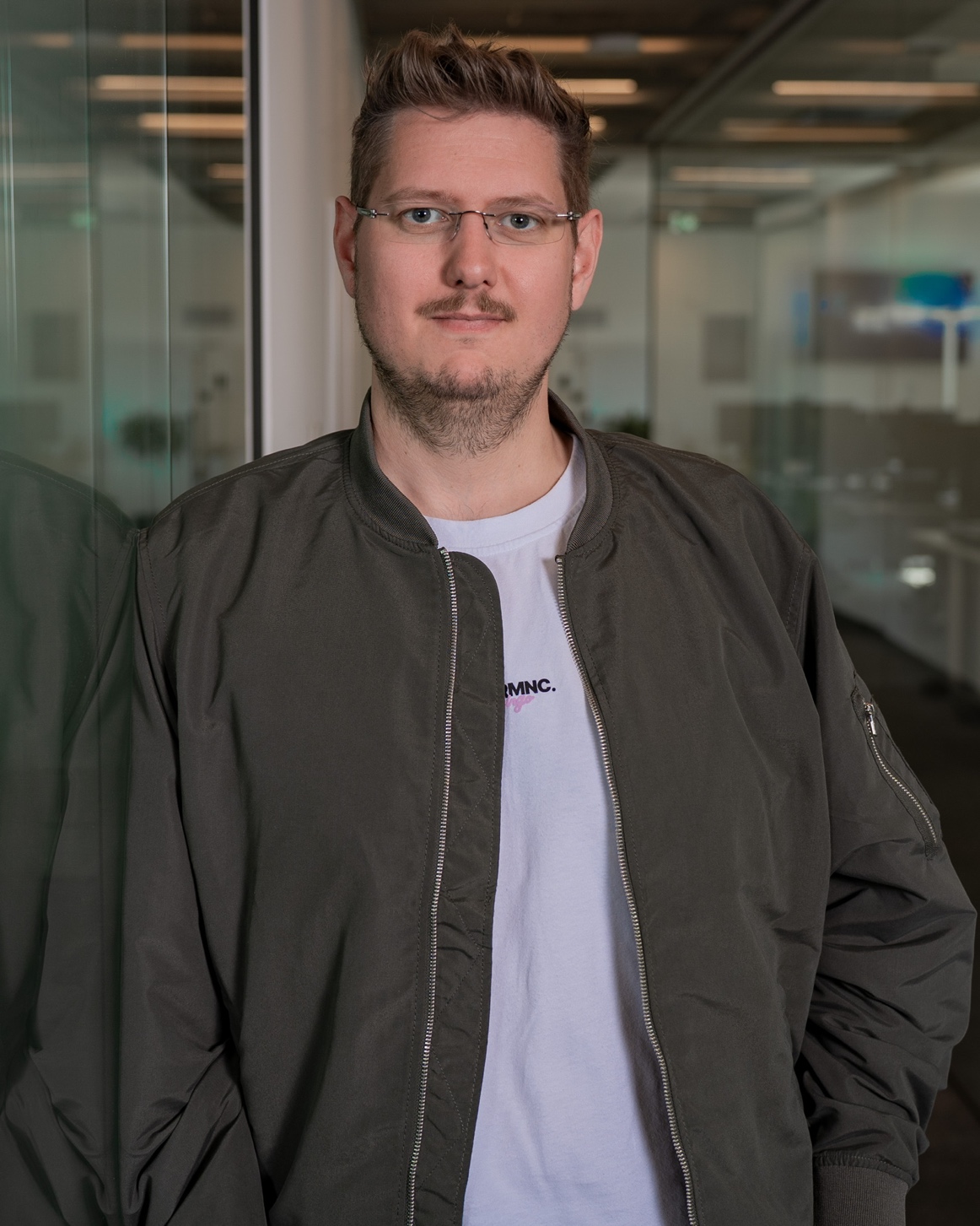 Ein Mann mit Brille und Bart trägt eine olivgrüne Jacke über einem weißen T-Shirt.