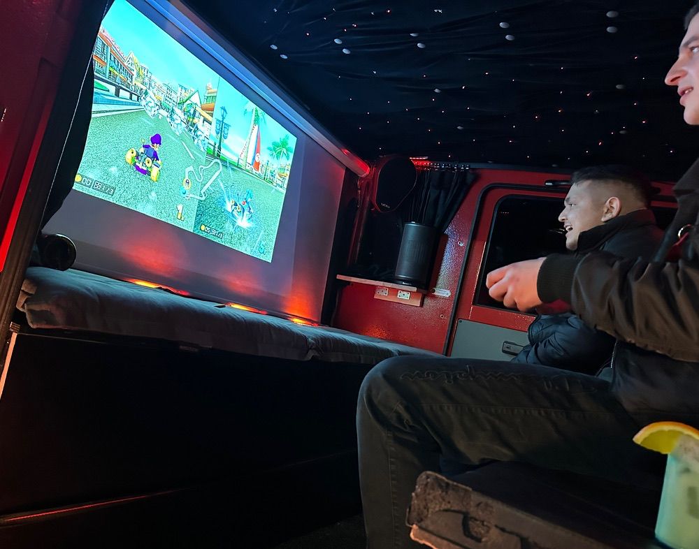 Zwei junge Männer spielen fröhlich Mario Kart auf einem großen Bildschirm im Van